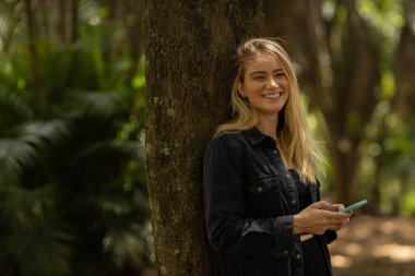 Gündüz vakti arka planda yeşil park olan akıllı telefon kullanan genç bir kadın. Yüksek kalite fotoğraf. Cep telefonu, teknoloji, şehir konsepti. Yüksek kalite fotoğraf