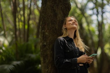 Gündüz vakti arka planda yeşil park olan akıllı telefon kullanan genç bir kadın. Yüksek kalite fotoğraf. Cep telefonu, teknoloji, şehir konsepti. Yüksek kalite fotoğraf