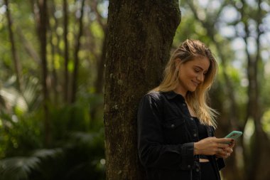 Gündüz vakti arka planda yeşil park olan akıllı telefon kullanan genç bir kadın. Yüksek kalite fotoğraf. Cep telefonu, teknoloji, şehir konsepti. Yüksek kalite fotoğraf