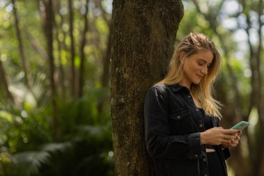 Gündüz vakti arka planda yeşil park olan akıllı telefon kullanan genç bir kadın. Yüksek kalite fotoğraf. Cep telefonu, teknoloji, şehir konsepti. Yüksek kalite fotoğraf