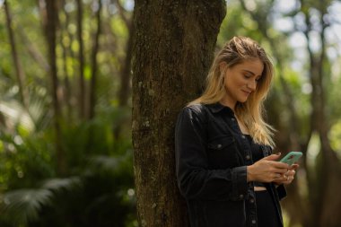 Gündüz vakti arka planda yeşil park olan akıllı telefon kullanan genç bir kadın. Yüksek kalite fotoğraf. Cep telefonu, teknoloji, şehir konsepti. Yüksek kalite fotoğraf