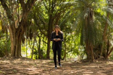 Gündüz vakti arka planda yeşil park olan akıllı telefon kullanan genç bir kadın. Yüksek kalite fotoğraf. Cep telefonu, teknoloji, şehir konsepti. Yüksek kalite fotoğraf