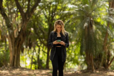 Gündüz vakti arka planda yeşil park olan akıllı telefon kullanan genç bir kadın. Yüksek kalite fotoğraf. Cep telefonu, teknoloji, şehir konsepti. Yüksek kalite fotoğraf