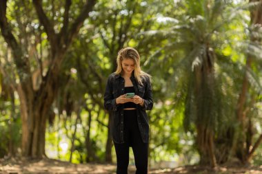 Gündüz vakti arka planda yeşil park olan akıllı telefon kullanan genç bir kadın. Yüksek kalite fotoğraf. Cep telefonu, teknoloji, şehir konsepti. Yüksek kalite fotoğraf