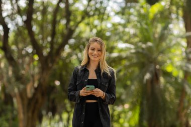 Gündüz vakti arka planda yeşil park olan akıllı telefon kullanan genç bir kadın. Yüksek kalite fotoğraf. Cep telefonu, teknoloji, şehir konsepti. Yüksek kalite fotoğraf