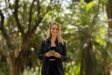 Gündüz vakti arka planda yeşil park olan akıllı telefon kullanan genç bir kadın. Yüksek kalite fotoğraf. Cep telefonu, teknoloji, şehir konsepti. Yüksek kalite fotoğraf