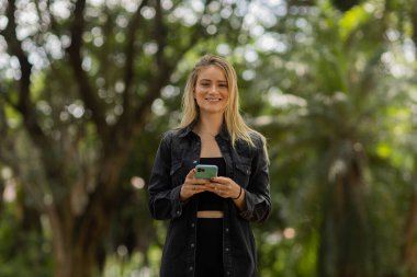 Gündüz vakti arka planda yeşil park olan akıllı telefon kullanan genç bir kadın. Yüksek kalite fotoğraf. Cep telefonu, teknoloji, şehir konsepti. Yüksek kalite fotoğraf