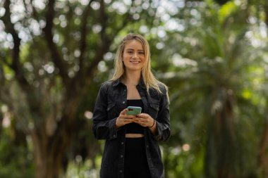 Gündüz vakti arka planda yeşil park olan akıllı telefon kullanan genç bir kadın. Yüksek kalite fotoğraf. Cep telefonu, teknoloji, şehir konsepti. Yüksek kalite fotoğraf