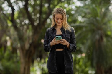 Gündüz vakti arka planda yeşil park olan akıllı telefon kullanan genç bir kadın. Yüksek kalite fotoğraf. Cep telefonu, teknoloji, şehir konsepti. Yüksek kalite fotoğraf