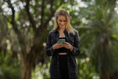 Gündüz vakti arka planda yeşil park olan akıllı telefon kullanan genç bir kadın. Yüksek kalite fotoğraf. Cep telefonu, teknoloji, şehir konsepti. Yüksek kalite fotoğraf