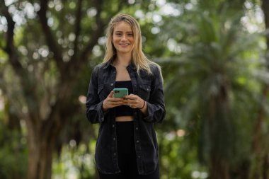 Gündüz vakti arka planda yeşil park olan akıllı telefon kullanan genç bir kadın. Yüksek kalite fotoğraf. Cep telefonu, teknoloji, şehir konsepti. Yüksek kalite fotoğraf