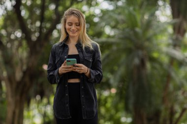 Gündüz vakti arka planda yeşil park olan akıllı telefon kullanan genç bir kadın. Yüksek kalite fotoğraf. Cep telefonu, teknoloji, şehir konsepti. Yüksek kalite fotoğraf