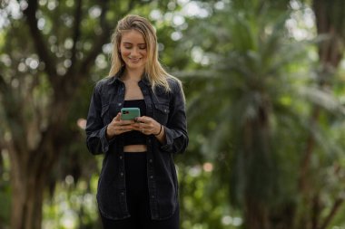 Gündüz vakti arka planda yeşil park olan akıllı telefon kullanan genç bir kadın. Yüksek kalite fotoğraf. Cep telefonu, teknoloji, şehir konsepti. Yüksek kalite fotoğraf