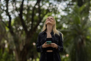 Gündüz vakti arka planda yeşil park olan akıllı telefon kullanan genç bir kadın. Yüksek kalite fotoğraf. Cep telefonu, teknoloji, şehir konsepti. Yüksek kalite fotoğraf