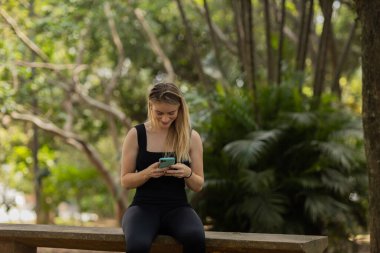 Gündüz vakti arka planda yeşil park olan akıllı telefon kullanan genç bir kadın. Yüksek kalite fotoğraf. Cep telefonu, teknoloji, şehir konsepti. Yüksek kalite fotoğraf