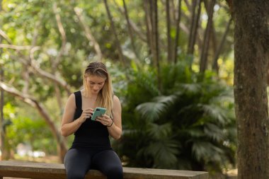 Gündüz vakti arka planda yeşil park olan akıllı telefon kullanan genç bir kadın. Yüksek kalite fotoğraf. Cep telefonu, teknoloji, şehir konsepti. Yüksek kalite fotoğraf