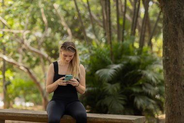 Gündüz vakti arka planda yeşil park olan akıllı telefon kullanan genç bir kadın. Yüksek kalite fotoğraf. Cep telefonu, teknoloji, şehir konsepti. Yüksek kalite fotoğraf