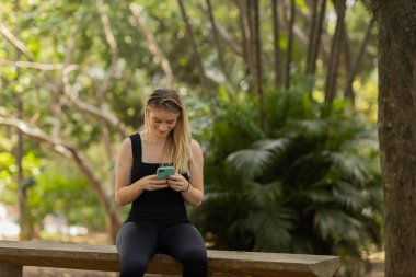 Gündüz vakti arka planda yeşil park olan akıllı telefon kullanan genç bir kadın. Yüksek kalite fotoğraf. Cep telefonu, teknoloji, şehir konsepti. Yüksek kalite fotoğraf