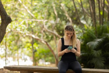Gündüz vakti arka planda yeşil park olan akıllı telefon kullanan genç bir kadın. Yüksek kalite fotoğraf. Cep telefonu, teknoloji, şehir konsepti. Yüksek kalite fotoğraf