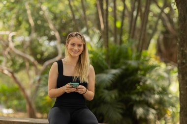 Gündüz vakti arka planda yeşil park olan akıllı telefon kullanan genç bir kadın. Yüksek kalite fotoğraf. Cep telefonu, teknoloji, şehir konsepti. Yüksek kalite fotoğraf
