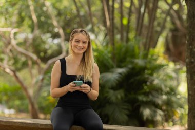 Gündüz vakti arka planda yeşil park olan akıllı telefon kullanan genç bir kadın. Yüksek kalite fotoğraf. Cep telefonu, teknoloji, şehir konsepti. Yüksek kalite fotoğraf
