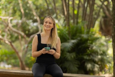 Gündüz vakti arka planda yeşil park olan akıllı telefon kullanan genç bir kadın. Yüksek kalite fotoğraf. Cep telefonu, teknoloji, şehir konsepti. Yüksek kalite fotoğraf