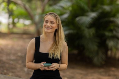 Gündüz vakti arka planda yeşil park olan akıllı telefon kullanan genç bir kadın. Yüksek kalite fotoğraf. Cep telefonu, teknoloji, şehir konsepti. Yüksek kalite fotoğraf