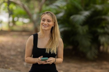Gündüz vakti arka planda yeşil park olan akıllı telefon kullanan genç bir kadın. Yüksek kalite fotoğraf. Cep telefonu, teknoloji, şehir konsepti. Yüksek kalite fotoğraf