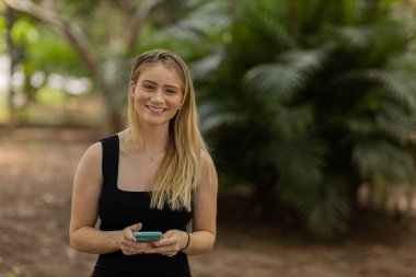 Gündüz vakti arka planda yeşil park olan akıllı telefon kullanan genç bir kadın. Yüksek kalite fotoğraf. Cep telefonu, teknoloji, şehir konsepti. Yüksek kalite fotoğraf