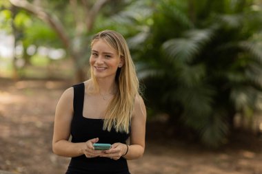 Gündüz vakti arka planda yeşil park olan akıllı telefon kullanan genç bir kadın. Yüksek kalite fotoğraf. Cep telefonu, teknoloji, şehir konsepti. Yüksek kalite fotoğraf