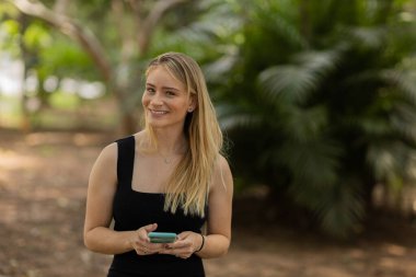Gündüz vakti arka planda yeşil park olan akıllı telefon kullanan genç bir kadın. Yüksek kalite fotoğraf. Cep telefonu, teknoloji, şehir konsepti. Yüksek kalite fotoğraf