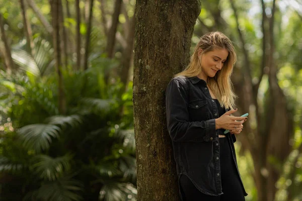 Gündüz vakti arka planda yeşil park olan akıllı telefon kullanan genç bir kadın. Yüksek kalite fotoğraf. Cep telefonu, teknoloji, şehir konsepti. Yüksek kalite fotoğraf