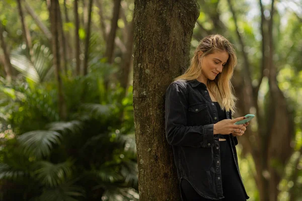 Gündüz vakti arka planda yeşil park olan akıllı telefon kullanan genç bir kadın. Yüksek kalite fotoğraf. Cep telefonu, teknoloji, şehir konsepti. Yüksek kalite fotoğraf
