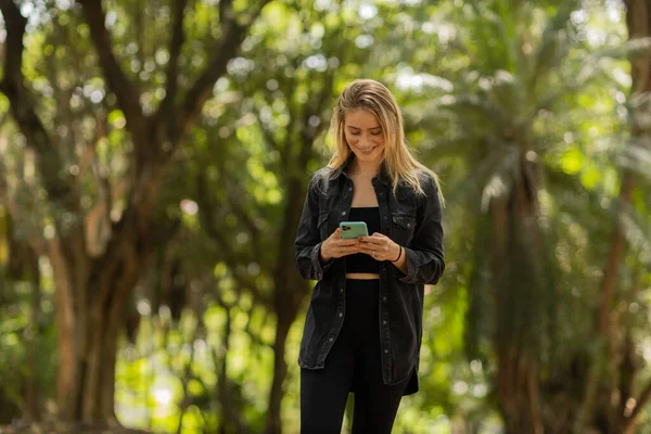 Gündüz vakti arka planda yeşil park olan akıllı telefon kullanan genç bir kadın. Yüksek kalite fotoğraf. Cep telefonu, teknoloji, şehir konsepti. Yüksek kalite fotoğraf