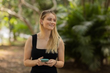 Gündüz vakti arka planda yeşil park olan akıllı telefon kullanan genç bir kadın. Yüksek kalite fotoğraf. Cep telefonu, teknoloji, şehir konsepti. Yüksek kalite fotoğraf