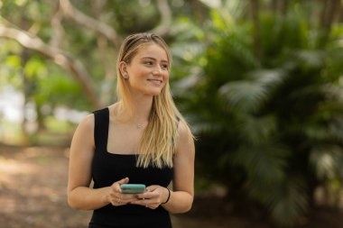 Gündüz vakti arka planda yeşil park olan akıllı telefon kullanan genç bir kadın. Yüksek kalite fotoğraf. Cep telefonu, teknoloji, şehir konsepti. Yüksek kalite fotoğraf
