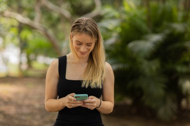 Gündüz vakti arka planda yeşil park olan akıllı telefon kullanan genç bir kadın. Yüksek kalite fotoğraf. Cep telefonu, teknoloji, şehir konsepti. Yüksek kalite fotoğraf