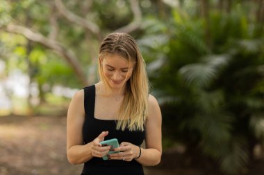 Gündüz vakti arka planda yeşil park olan akıllı telefon kullanan genç bir kadın. Yüksek kalite fotoğraf. Cep telefonu, teknoloji, şehir konsepti. Yüksek kalite fotoğraf