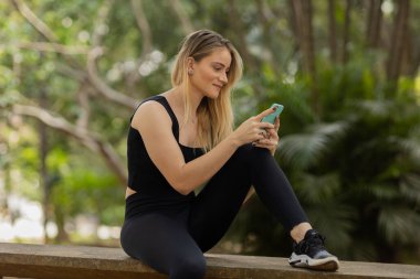 Gündüz vakti arka planda yeşil park olan akıllı telefon kullanan genç bir kadın. Yüksek kalite fotoğraf. Cep telefonu, teknoloji, şehir konsepti. Yüksek kalite fotoğraf
