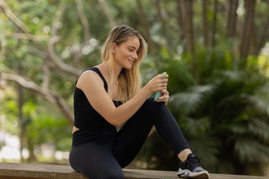 Gündüz vakti arka planda yeşil park olan akıllı telefon kullanan genç bir kadın. Yüksek kalite fotoğraf. Cep telefonu, teknoloji, şehir konsepti. Yüksek kalite fotoğraf