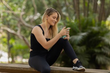 Gündüz vakti arka planda yeşil park olan akıllı telefon kullanan genç bir kadın. Yüksek kalite fotoğraf. Cep telefonu, teknoloji, şehir konsepti. Yüksek kalite fotoğraf
