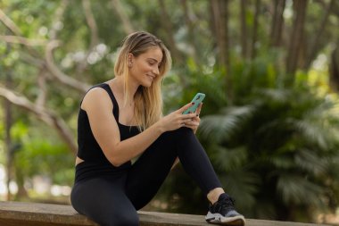 Gündüz vakti arka planda yeşil park olan akıllı telefon kullanan genç bir kadın. Yüksek kalite fotoğraf. Cep telefonu, teknoloji, şehir konsepti. Yüksek kalite fotoğraf