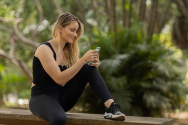 Gündüz vakti arka planda yeşil park olan akıllı telefon kullanan genç bir kadın. Yüksek kalite fotoğraf. Cep telefonu, teknoloji, şehir konsepti. Yüksek kalite fotoğraf