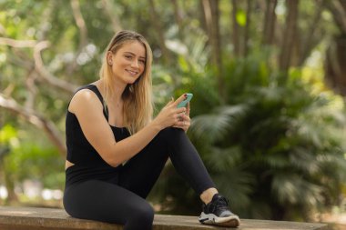 Gündüz vakti arka planda yeşil park olan akıllı telefon kullanan genç bir kadın. Yüksek kalite fotoğraf. Cep telefonu, teknoloji, şehir konsepti. Yüksek kalite fotoğraf