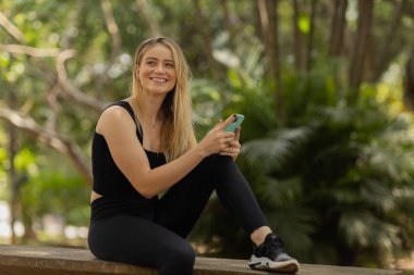 Gündüz vakti arka planda yeşil park olan akıllı telefon kullanan genç bir kadın. Yüksek kalite fotoğraf. Cep telefonu, teknoloji, şehir konsepti. Yüksek kalite fotoğraf