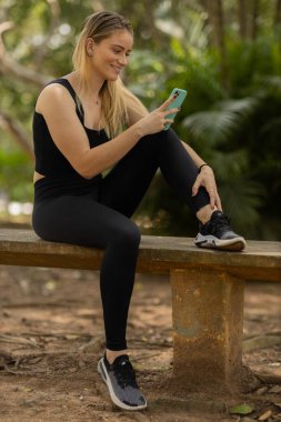 Gündüz vakti arka planda yeşil park olan akıllı telefon kullanan genç bir kadın. Yüksek kalite fotoğraf. Cep telefonu, teknoloji, şehir konsepti. Yüksek kalite fotoğraf