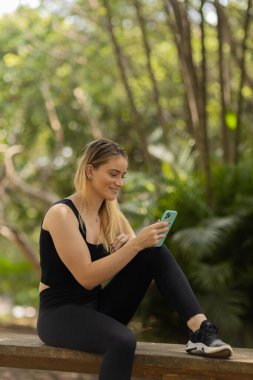 Gündüz vakti arka planda yeşil park olan akıllı telefon kullanan genç bir kadın. Yüksek kalite fotoğraf. Cep telefonu, teknoloji, şehir konsepti. Yüksek kalite fotoğraf