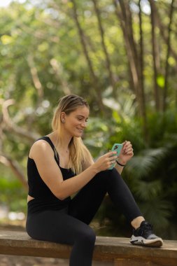 Gündüz vakti arka planda yeşil park olan akıllı telefon kullanan genç bir kadın. Yüksek kalite fotoğraf. Cep telefonu, teknoloji, şehir konsepti. Yüksek kalite fotoğraf