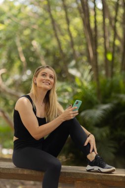 Gündüz vakti arka planda yeşil park olan akıllı telefon kullanan genç bir kadın. Yüksek kalite fotoğraf. Cep telefonu, teknoloji, şehir konsepti. Yüksek kalite fotoğraf