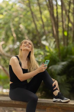Gündüz vakti arka planda yeşil park olan akıllı telefon kullanan genç bir kadın. Yüksek kalite fotoğraf. Cep telefonu, teknoloji, şehir konsepti. Yüksek kalite fotoğraf